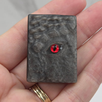 A small rectangular dark gray stone or resin pendant with a textured surface and a central red eye detail, held in a person's hand.