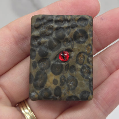 A hand holding a small rectangular pendant with a textured, organic dark pattern and a small red heart in the center, set against a light marble background.