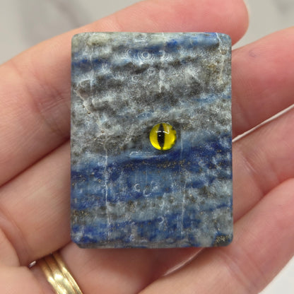 A hand holding a small rectangular blue and gray stone tile with a central yellow eye motif, shown against a light marble-like background.