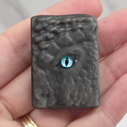 A small rectangular dark gray textured item with a blue eye detail, held in a person's hand against a light marble background.