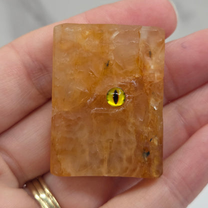 A hand holding a small rectangular amber-brown stone or resin piece with a small yellow-and-black eye charm in the center, against a light marble-like background.