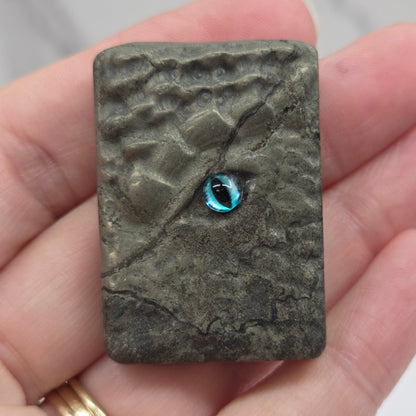 A hand holding a textured rectangular stone or ceramic tile with a small blue oval inlay near the center, against a light marble background.