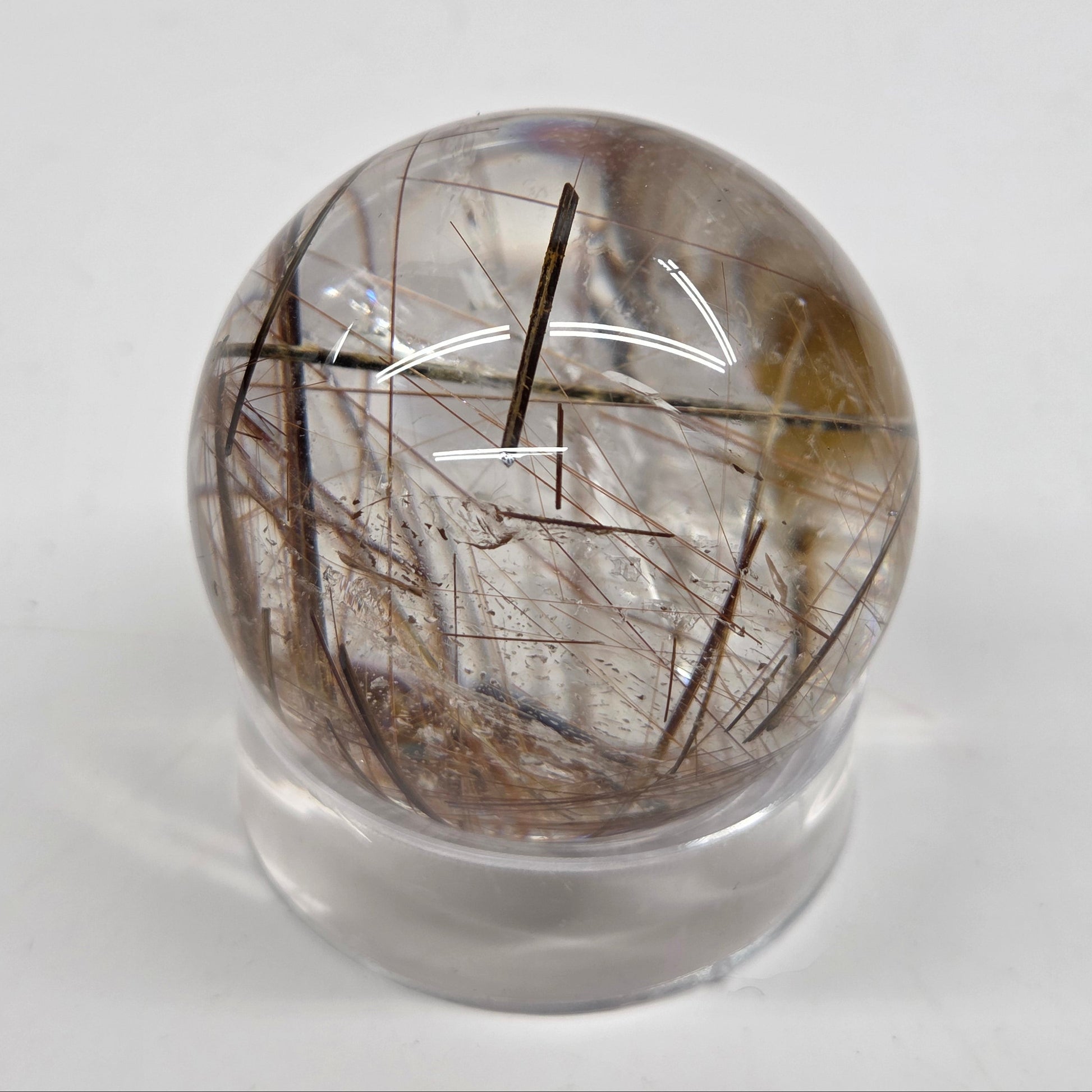 A clear quartz sphere containing rutile inclusions, displayed on a small clear stand against a plain light gray background.