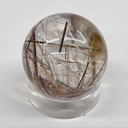 A clear quartz sphere containing rutile inclusions, displayed on a small clear stand against a plain light gray background.