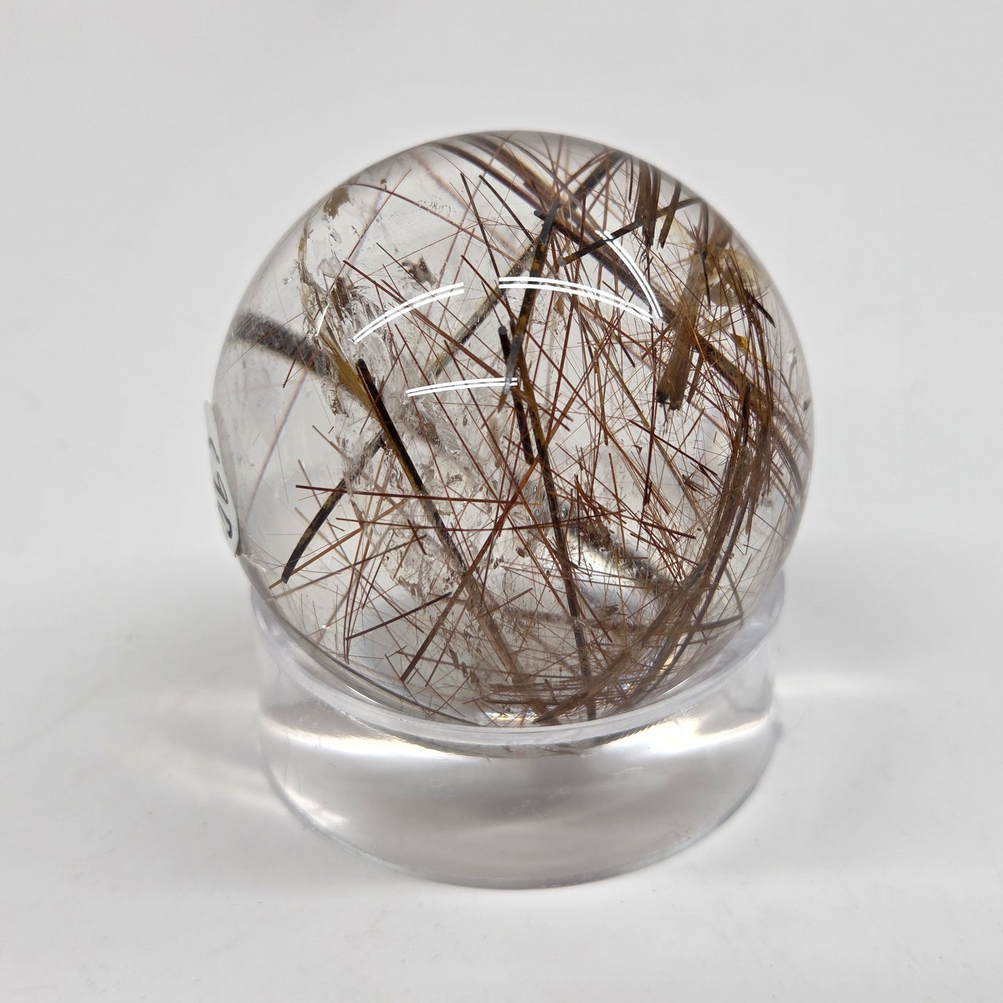 A clear glass or acrylic sphere containing numerous dark brown to black hair-like inclusions, displayed on a small clear stand against a light gray background.