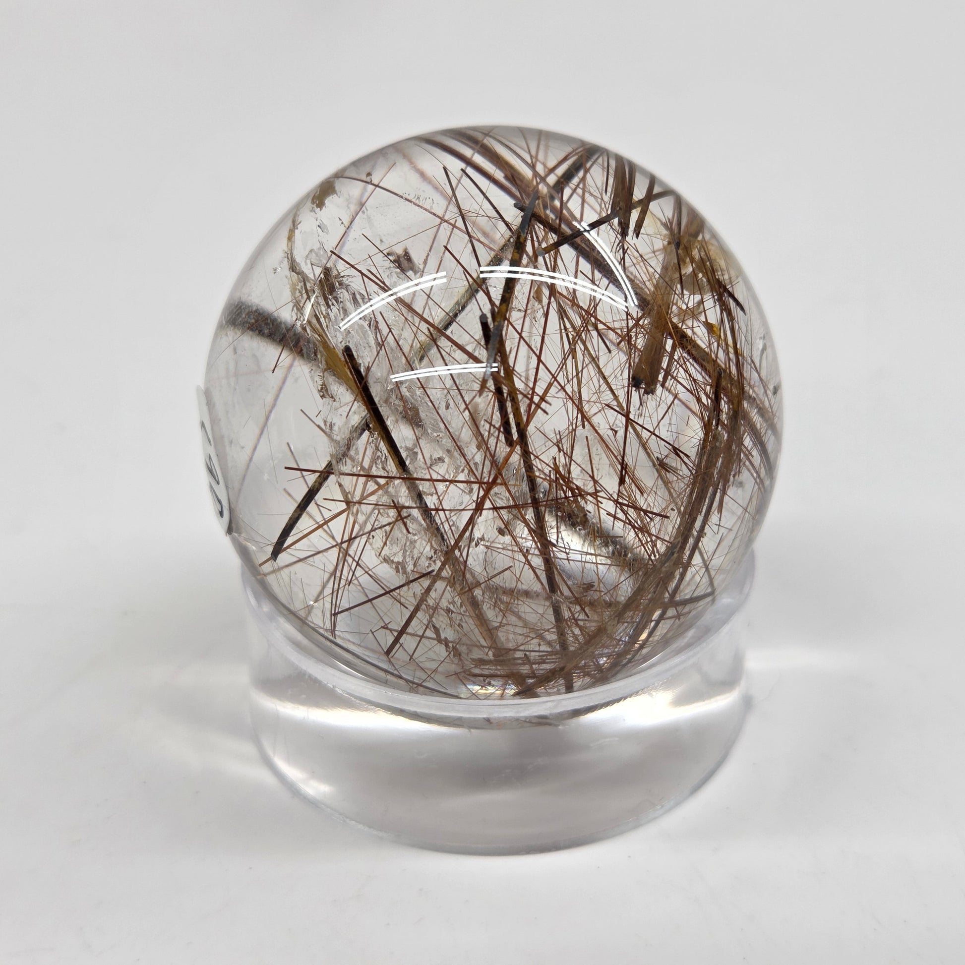 A clear glass or acrylic sphere containing numerous dark brown to black hair-like inclusions, displayed on a small clear stand against a light gray background.