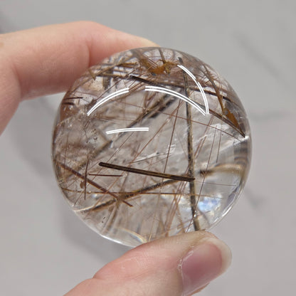 A hand holding a round clear crystal sphere containing fine brown and black needle-like inclusions, photographed against a light gray marble-like background.