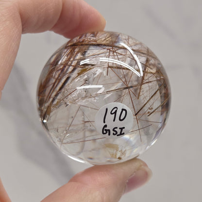 A hand holds a clear glass sphere containing rutile, with handwritten text reading '190 GSI' on the front, against a light gray marble-patterned background.