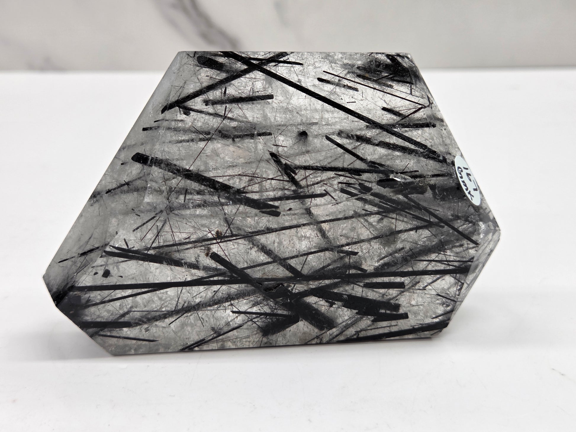 A faceted clear crystal point with dense black needle-like inclusions, photographed on a white surface against a light gray background.