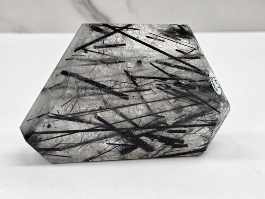 A faceted clear crystal point with dense black needle-like inclusions, photographed on a white surface against a light gray background.