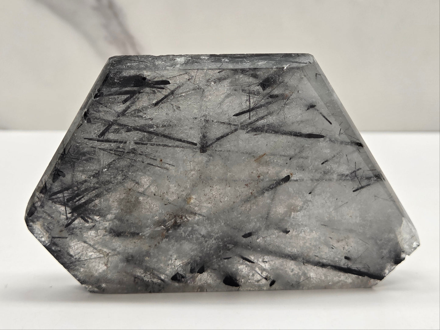 A faceted gray gemstone with prominent black needle-like inclusions, photographed on a white surface against a light gray background.