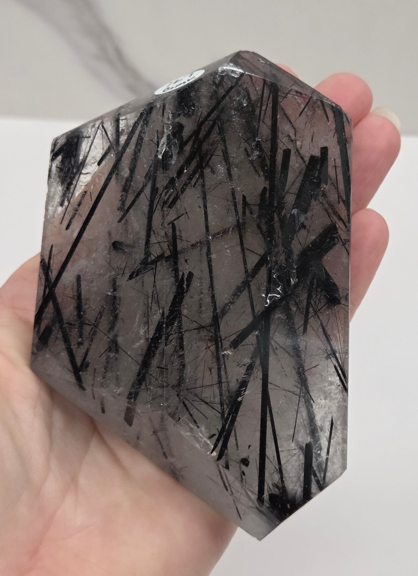 A hand holding a faceted clear crystal point with dense black needle-like inclusions, photographed against a light gray marble-patterned background.