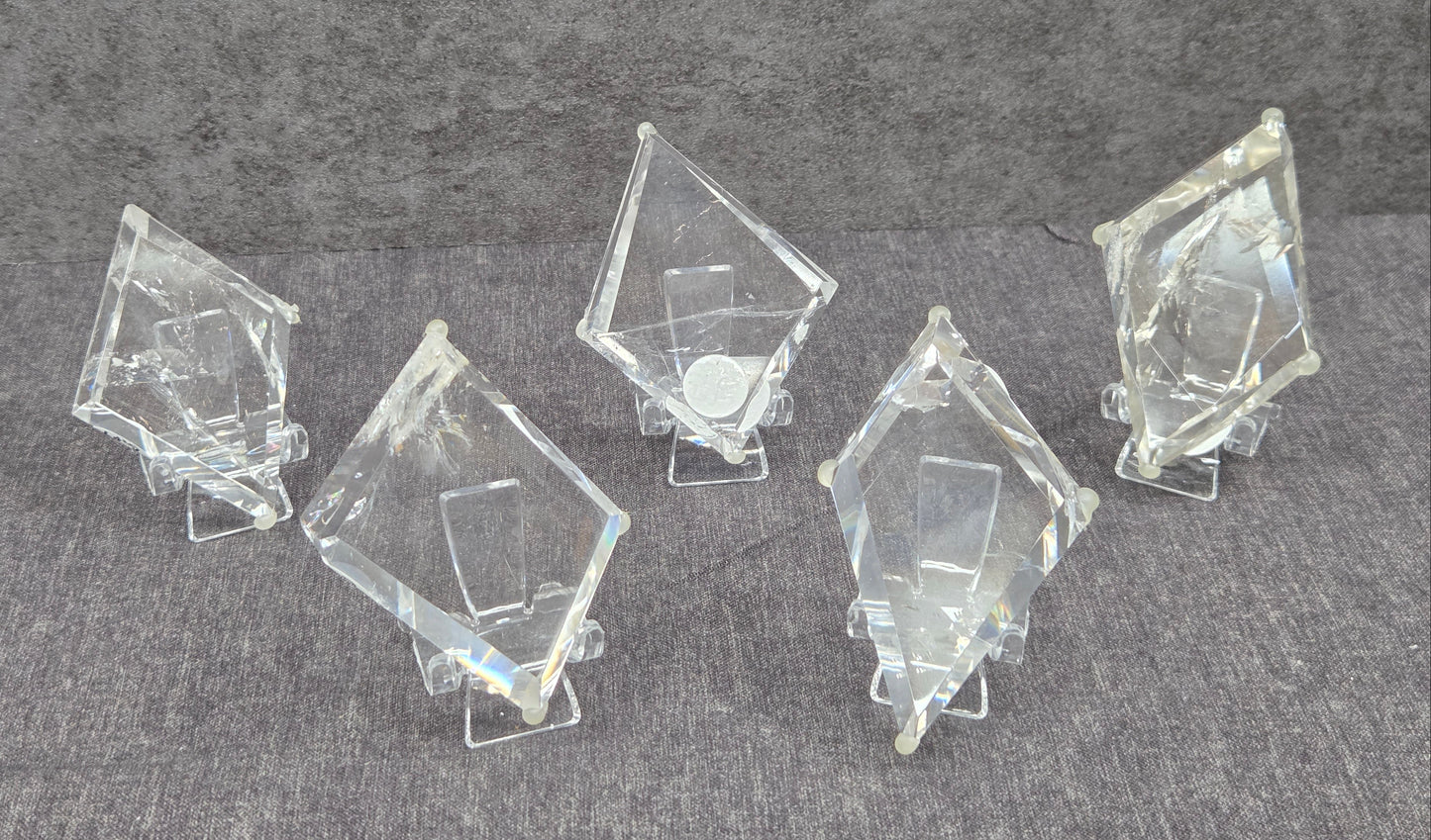 Five clear acrylic or glass geometric display blocks with triangular prismatic shapes, arranged on small clear stands against a textured gray background.