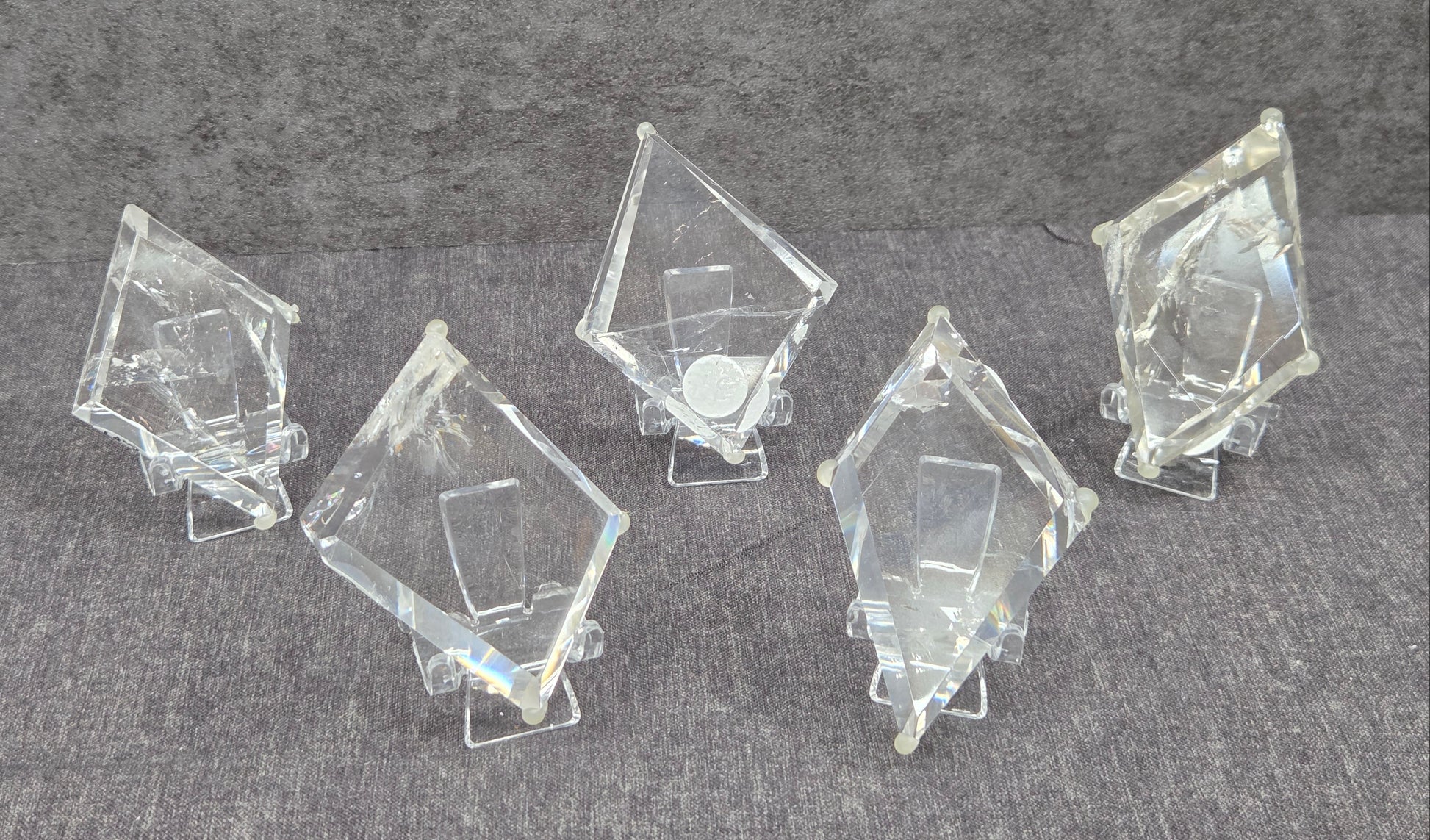 Five clear acrylic or glass geometric display blocks with triangular prismatic shapes, arranged on small clear stands against a textured gray background.