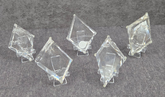 Five clear acrylic or glass geometric display blocks with triangular prismatic shapes, arranged on small clear stands against a textured gray background.