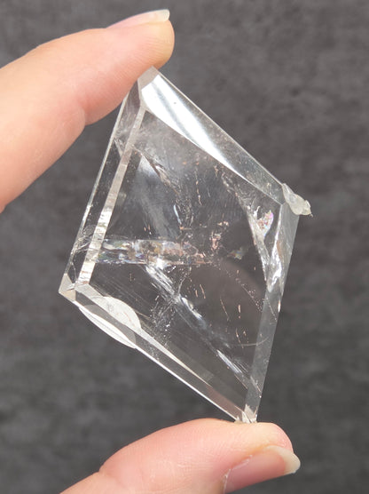A hand holding a clear, square glass or acrylic tile against a dark gray textured background.