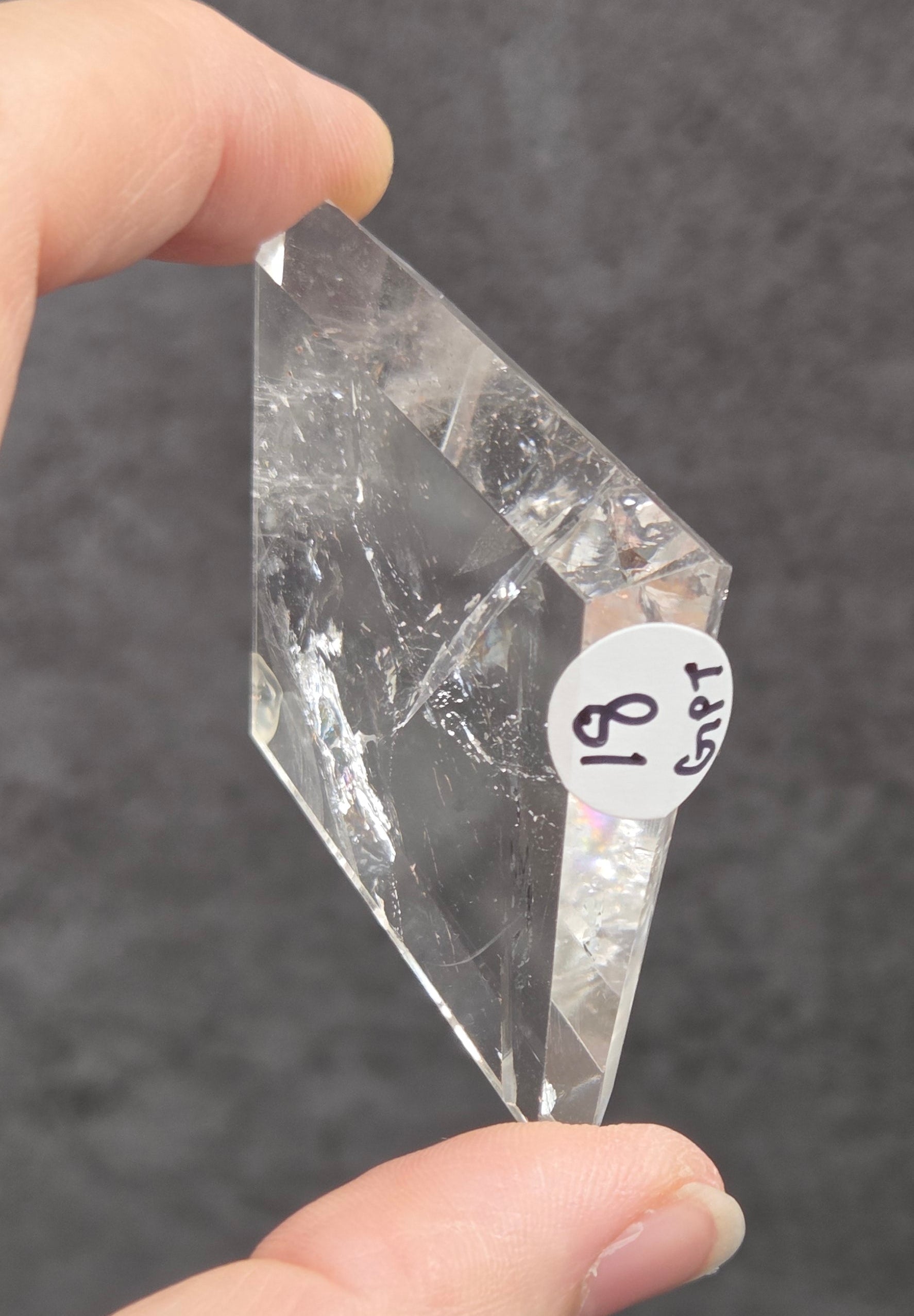 A hand holding a clear, faceted crystal or quartz point with a small white label that reads 'SRT 81' against a dark gray textured background.