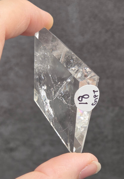 A hand holding a clear, faceted crystal or quartz point with a small white label that reads 'SRT 81' against a dark gray textured background.