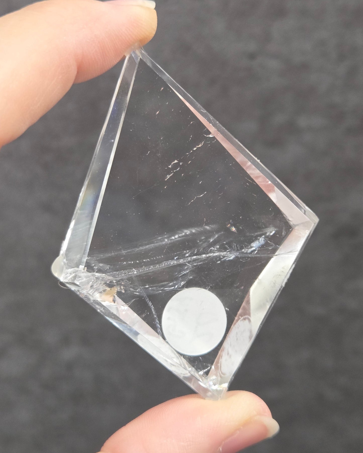 A hand holding a clear diamond-shaped acrylic block with a small white circular piece inside, set against a dark gray textured background.