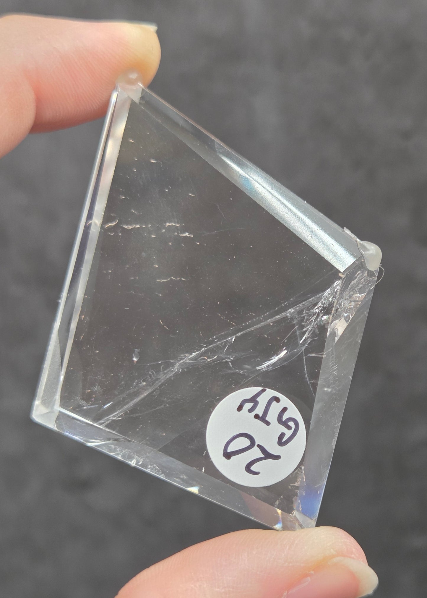 A hand holding a clear, transparent square glass block with a small white sticker labeled '20 G/TY' on it, set against a dark gray textured background.