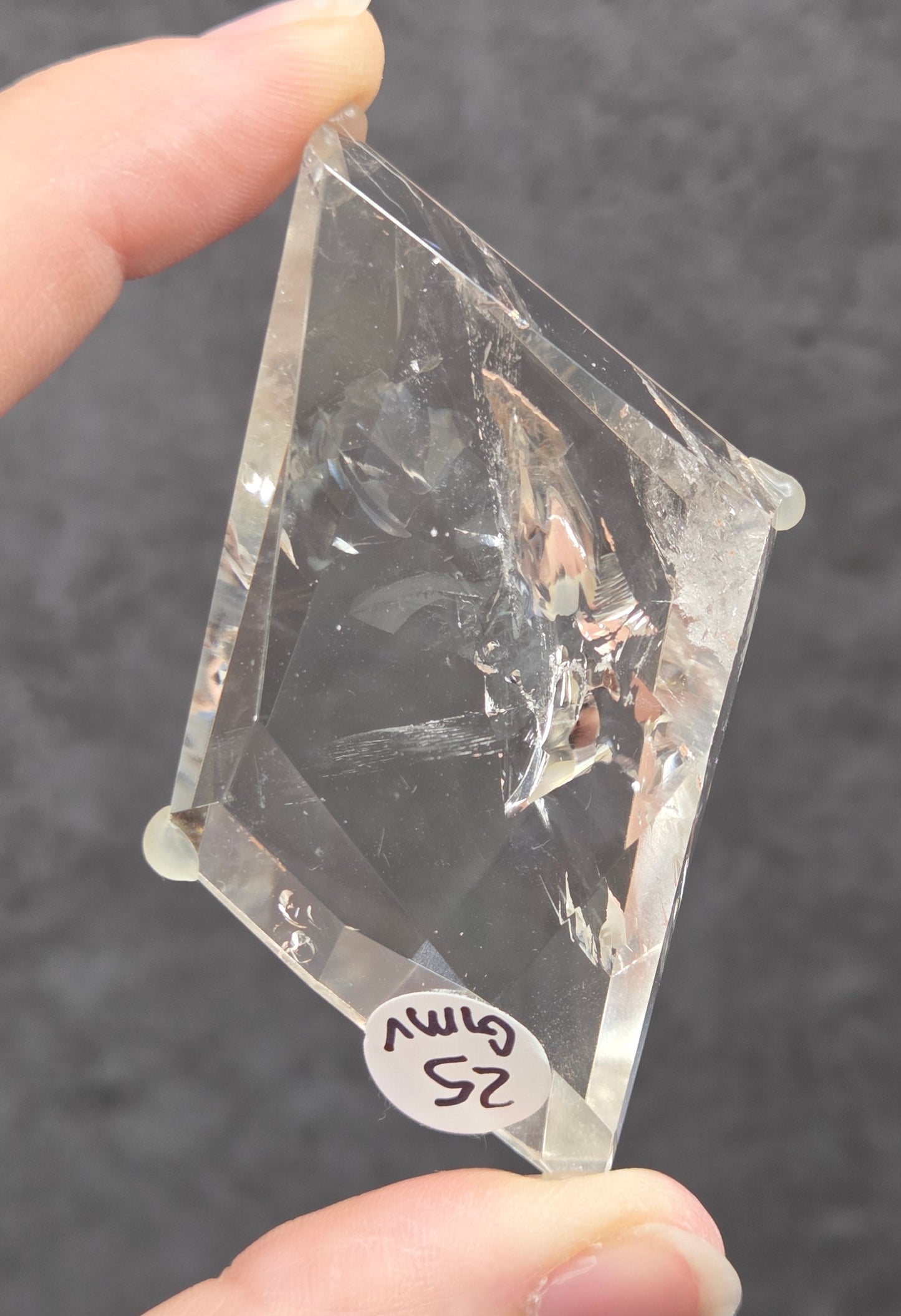 A hand holding a clear, diamond-shaped acrylic or glass specimen with a small white label attached, set against a dark textured gray background.