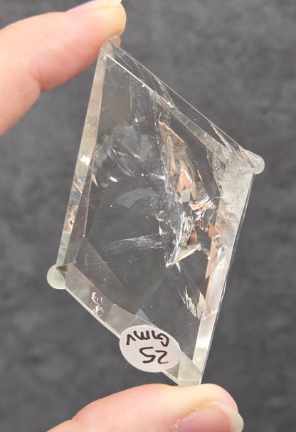 A hand holding a clear, diamond-shaped acrylic or glass specimen with a small white label attached, set against a dark textured gray background.