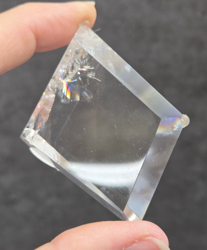 A hand holding a clear, faceted glass or acrylic cube against a dark gray textured background.