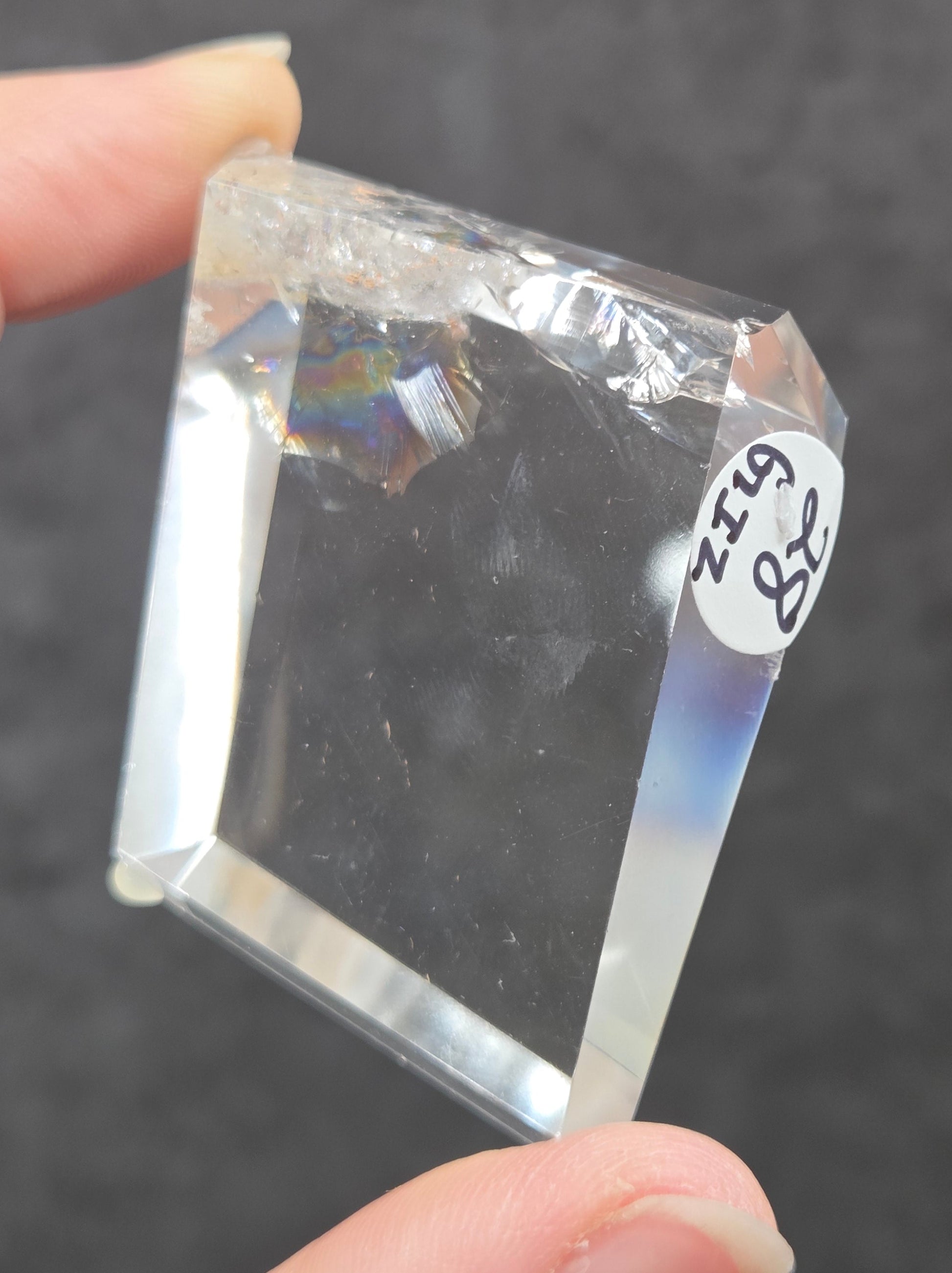 A hand holding a clear, faceted crystal or glass block with a small white label reading '48 Ginz' on it, against a dark gray textured background.