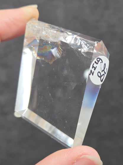 A hand holding a clear, faceted crystal or glass block with a small white label reading '48 Ginz' on it, against a dark gray textured background.