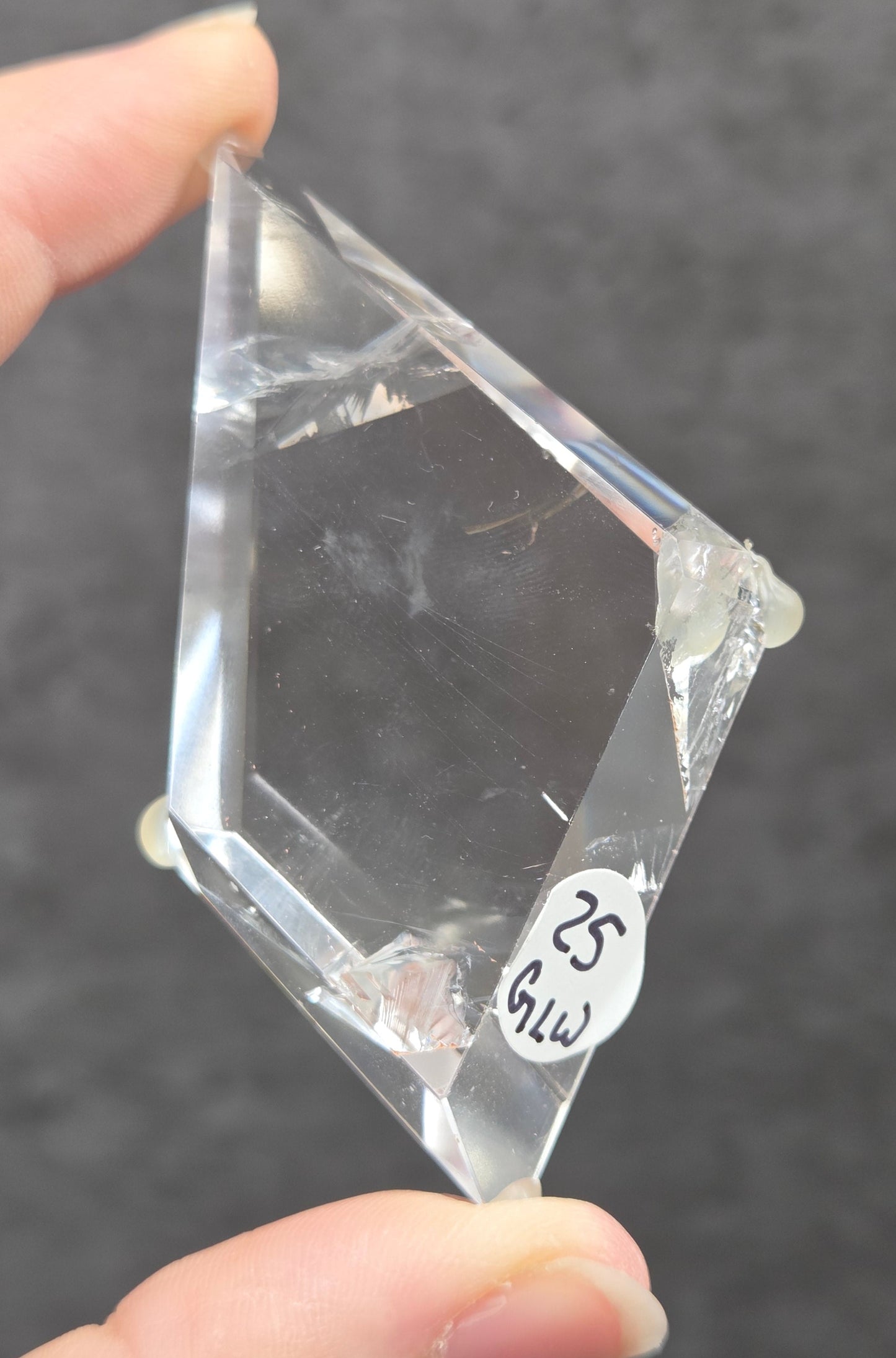 A hand holding a clear, faceted triangular glass prism against a dark gray textured background. A small white sticker with handwritten text is attached to one edge of the glass.