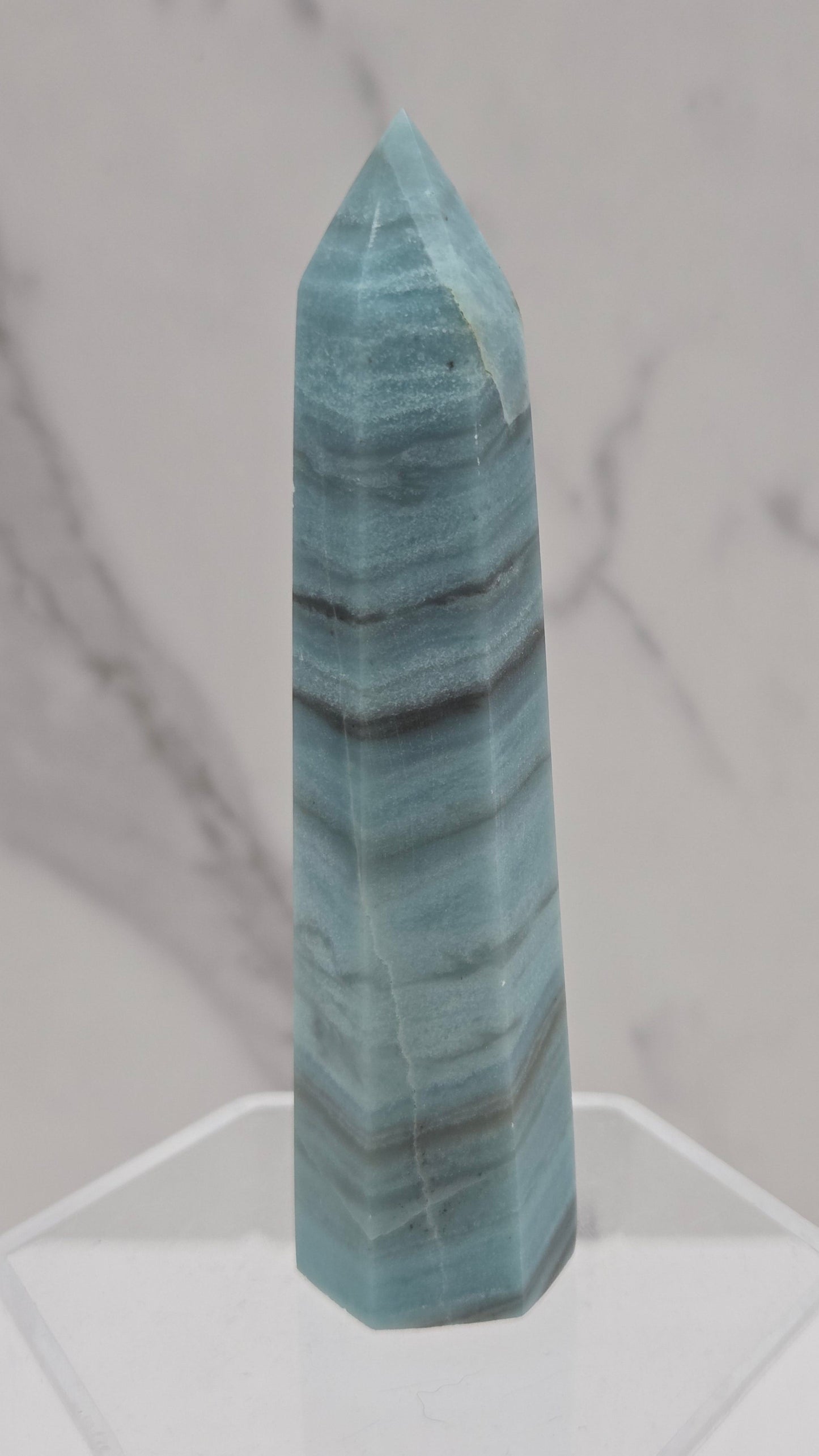 A polished blue-green stone crystal point with dark veining, displayed on a clear acrylic stand against a white marble background.