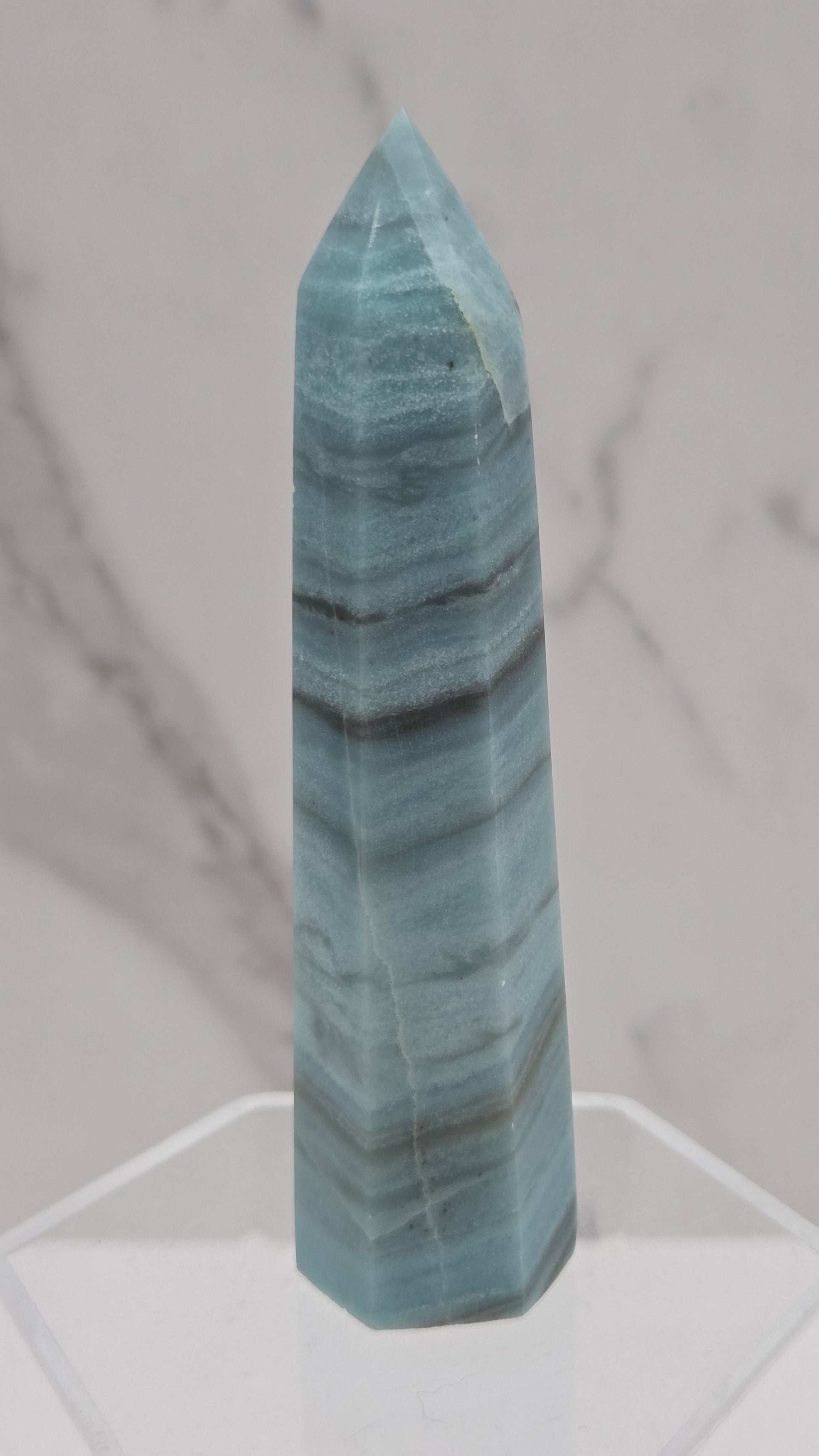 A polished blue-green stone crystal point with dark veining, displayed on a clear acrylic stand against a white marble background.