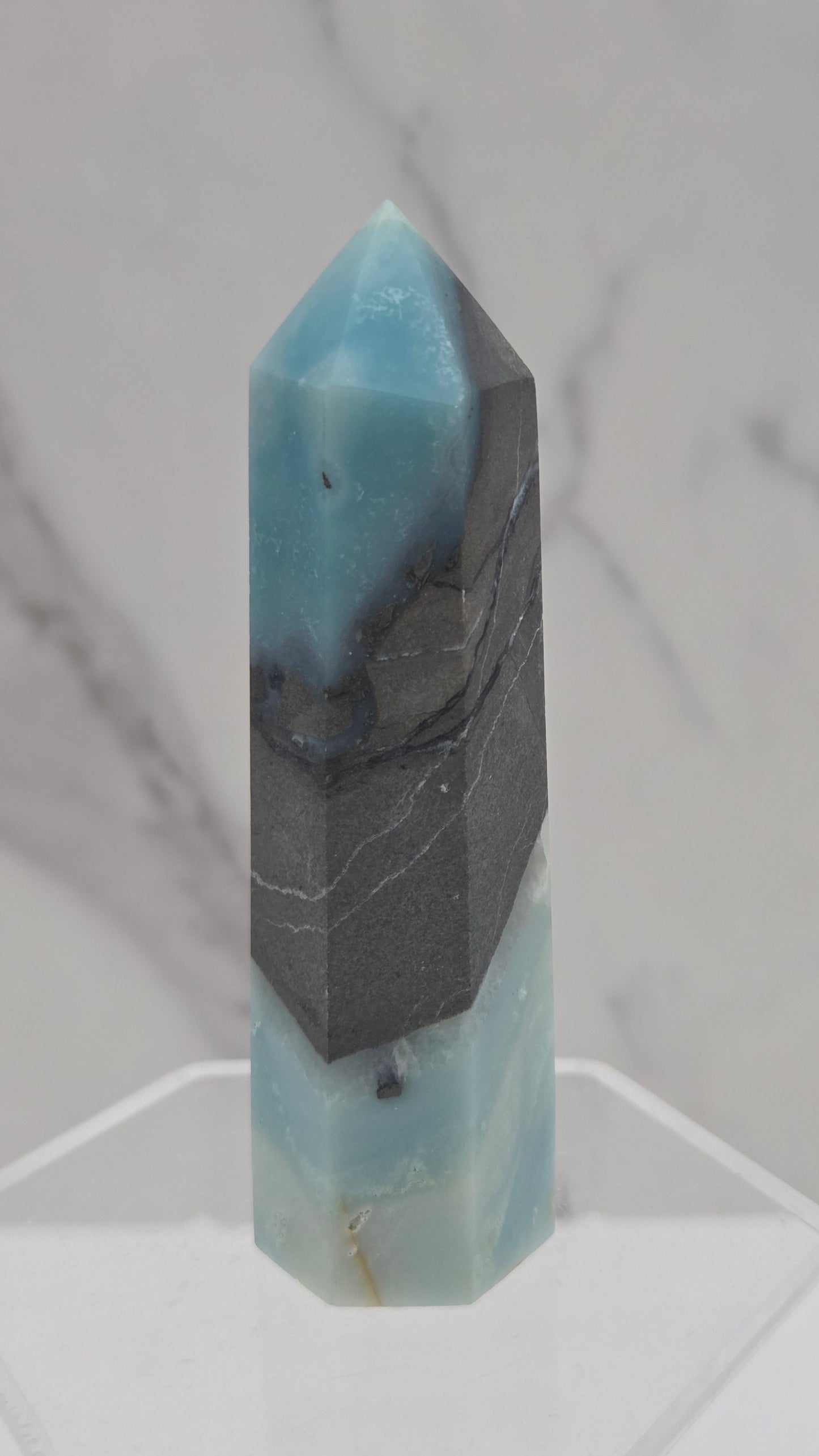 A polished calcite crystal point with a dark gray banded section, displayed on a clear acrylic stand against a white marble background.