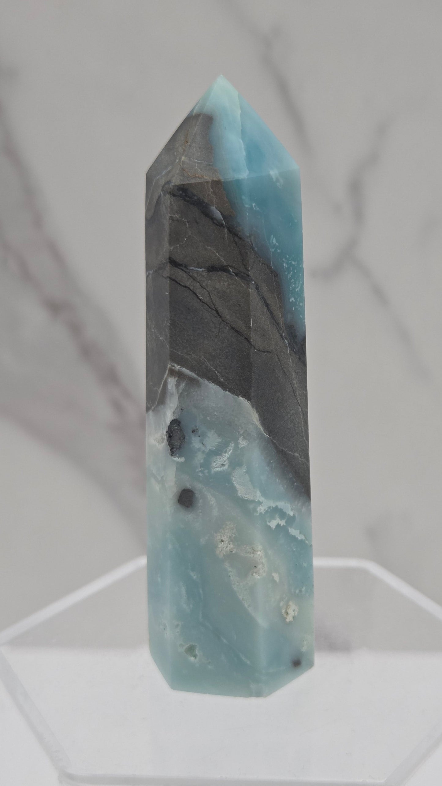 A polished calcite crystal point with natural blue and gray banded patterns, displayed on a clear acrylic stand against a white marble background.