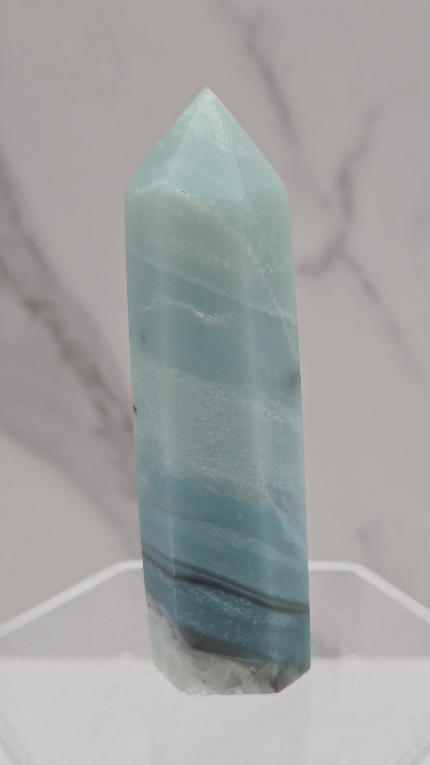 A polished light blue calcite crystal point displayed on a clear acrylic stand against a white and gray marble-patterned background.