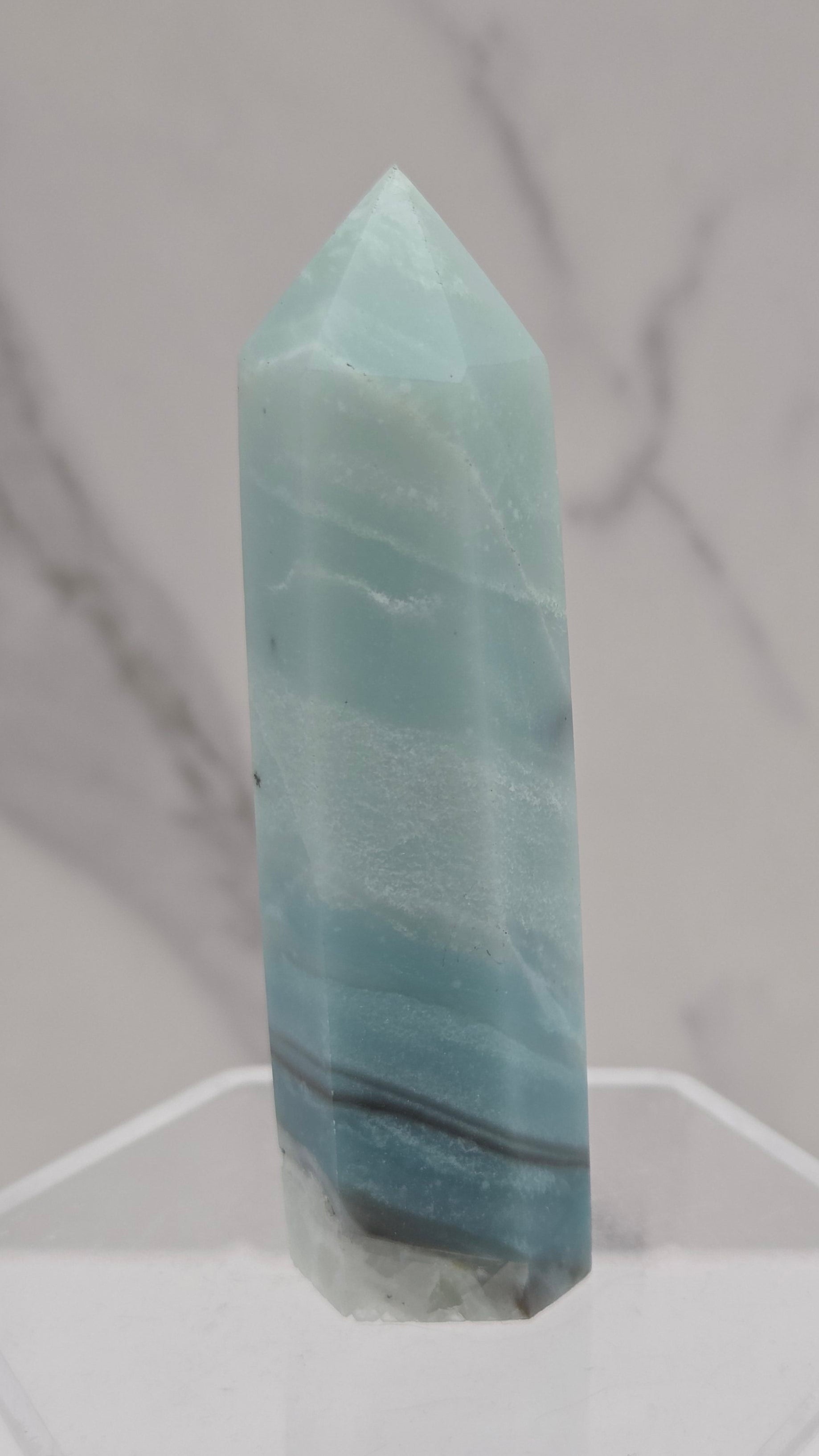 A polished light blue calcite crystal point displayed on a clear acrylic stand against a white and gray marble-patterned background.