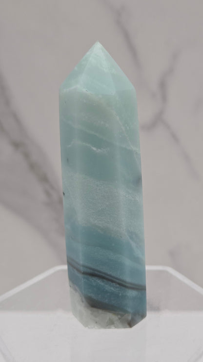 A polished light blue calcite crystal point displayed on a clear acrylic stand against a white and gray marble-patterned background.