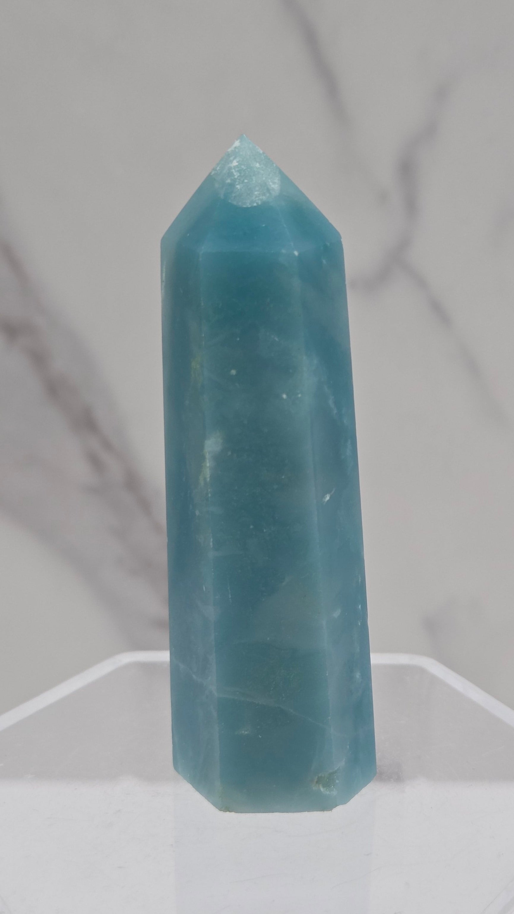 A light blue calcite crystal point displayed on a clear acrylic stand against a white and gray marble-patterned background.