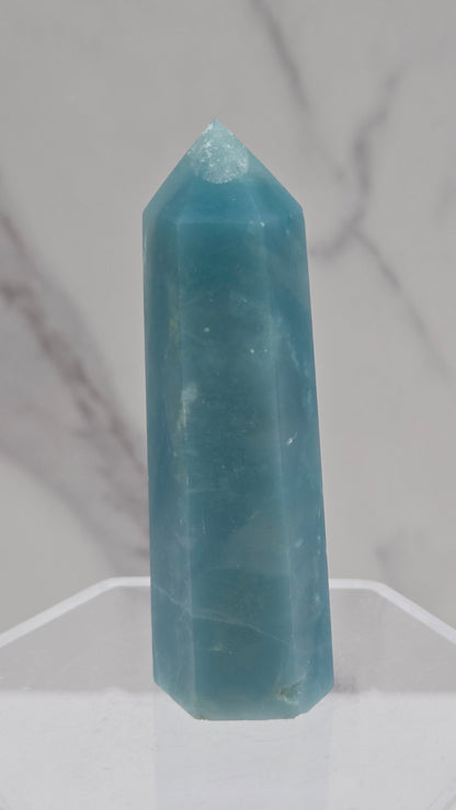 A light blue calcite crystal point displayed on a clear acrylic stand against a white and gray marble-patterned background.