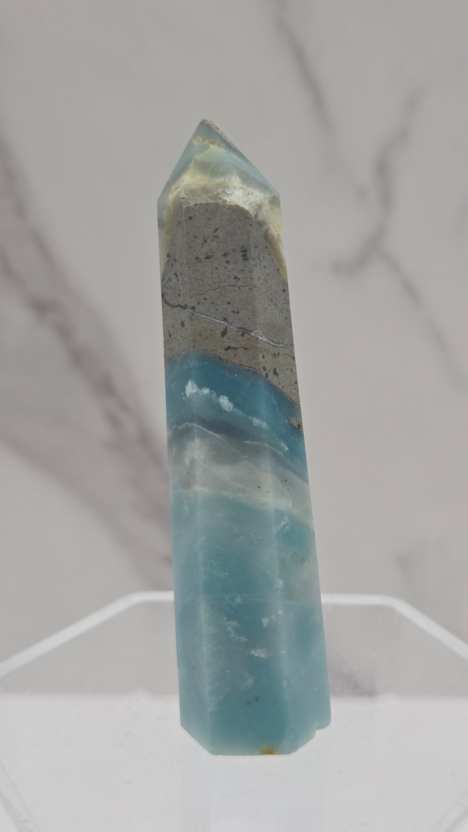 A polished blue and gray calcite crystal point displayed horizontally on a clear acrylic stand against a white marble background.