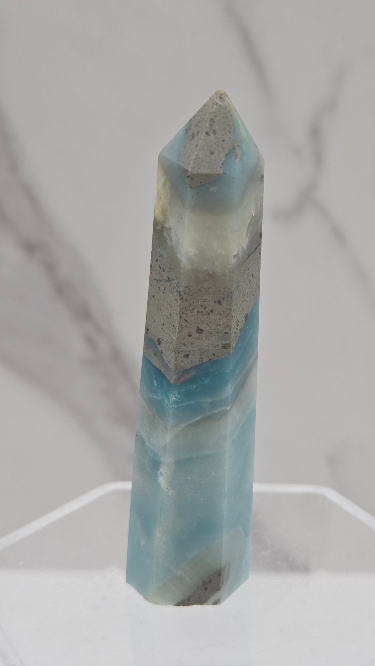 A polished calcite crystal point with light blue and gray marbling, displayed on a clear acrylic stand against a white marble background.