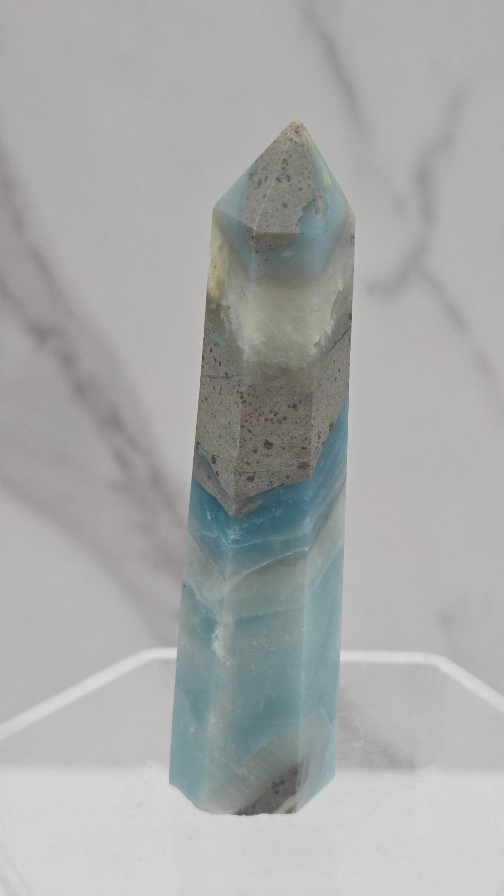A polished calcite crystal point with light blue and gray marbling, displayed on a clear acrylic stand against a white marble background.