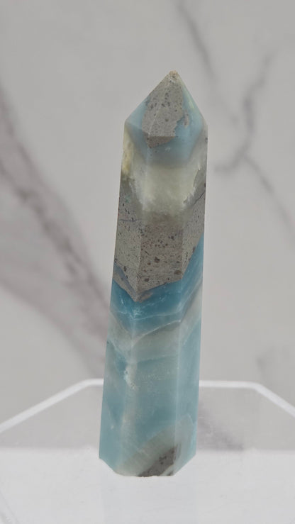 A polished calcite crystal point with light blue and gray marbling, displayed on a clear acrylic stand against a white marble background.