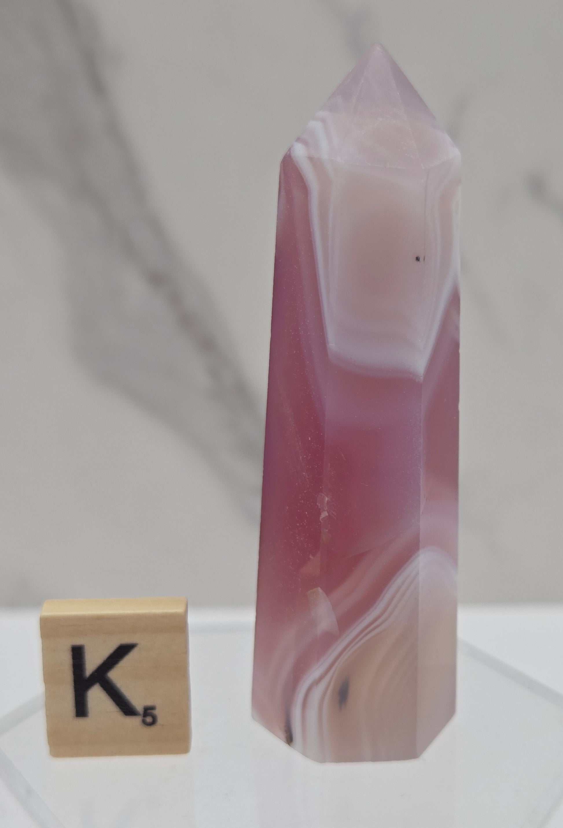 A polished pink and white agate crystal point displayed horizontally on a clear acrylic stand, with a wooden Scrabble tile showing the letter K below it against a light marble-patterned background.