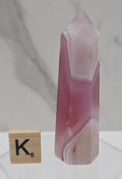 A polished pink and white agate crystal point displayed horizontally on a clear acrylic stand, with a wooden Scrabble tile showing the letter K below it against a light marble-patterned background.
