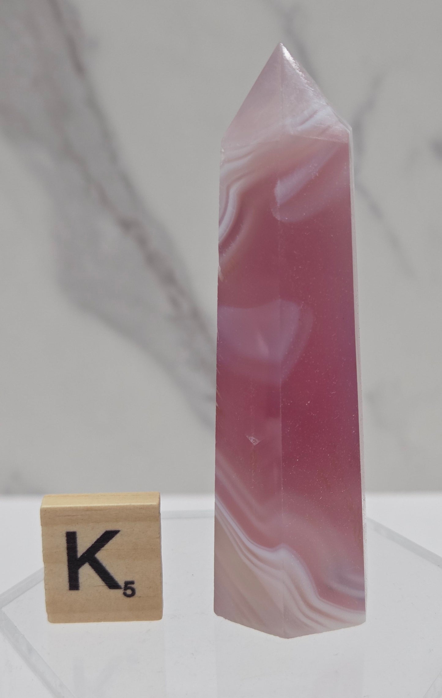 A polished pink agate crystal point with natural white veining, displayed horizontally on a clear acrylic stand next to a wooden Scrabble tile showing the letter K.
