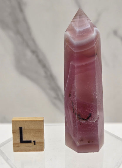 A faceted pinkish-red crystal point displayed horizontally on a clear acrylic stand, with a small wooden block showing the letter 'L' for scale, against a light marble-patterned background.