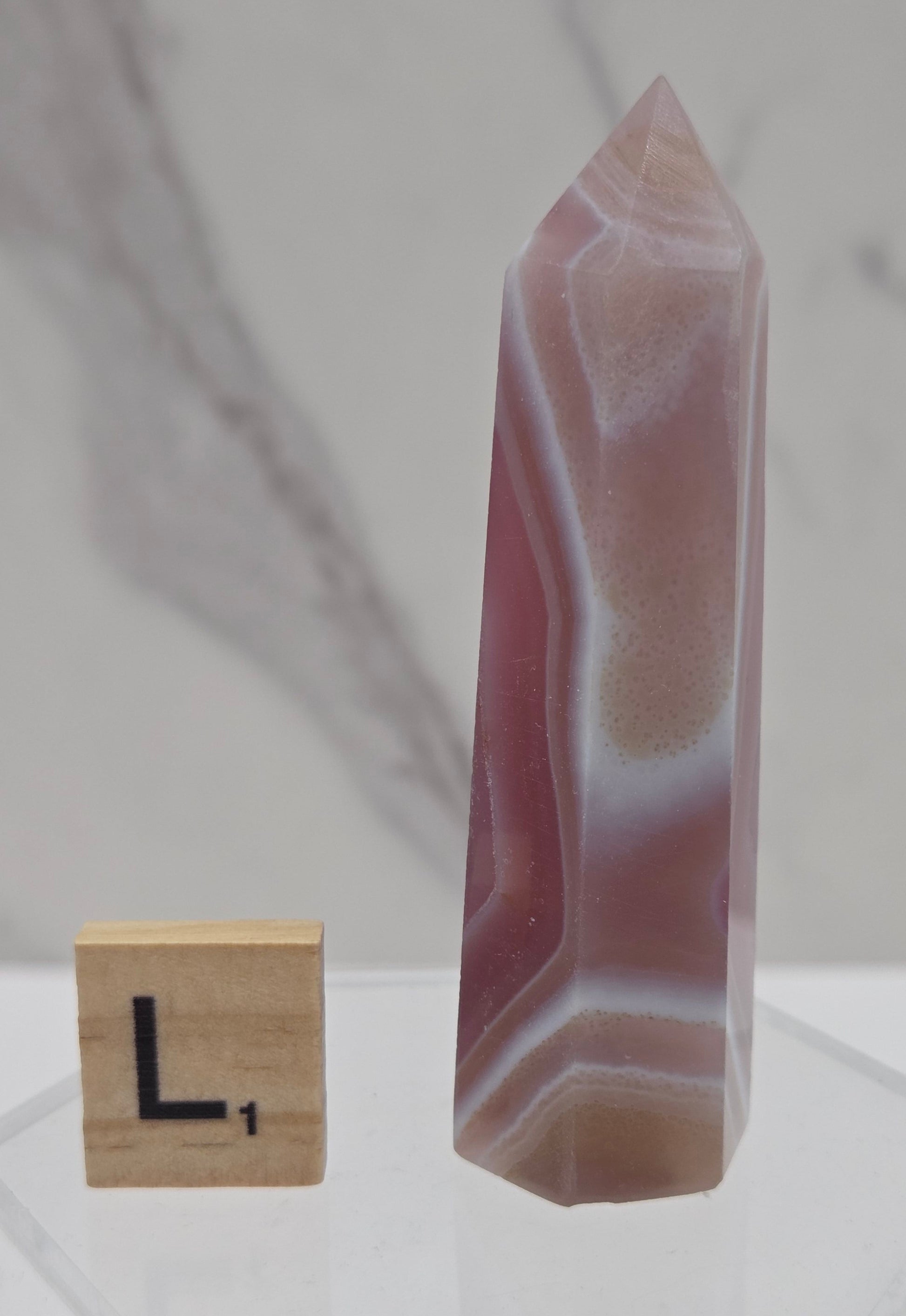 A polished agate crystal point with natural banding in pink, white, and brown, displayed horizontally next to a small wooden block on a clear acrylic stand against a light marble-patterned background.