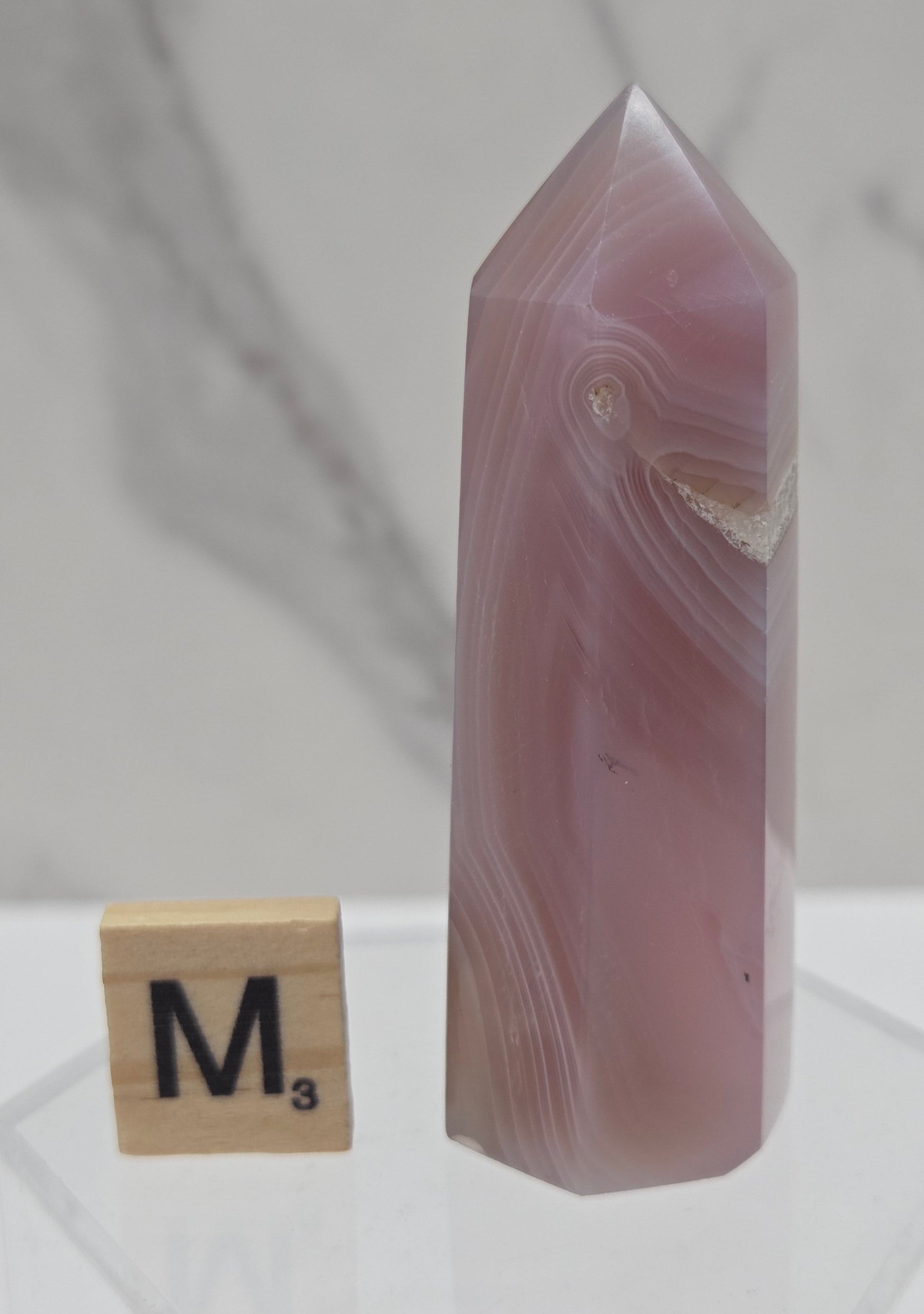 A polished pink agate crystal point displayed on a clear acrylic stand next to a wooden Scrabble tile marked 'M' on a light marble surface.