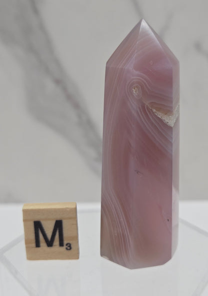 A polished pink agate crystal point displayed on a clear acrylic stand next to a wooden Scrabble tile marked 'M' on a light marble surface.